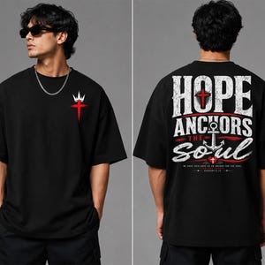 May include: A black short-sleeve t-shirt featuring a small red cross and white crown graphic on the front left chest. The back displays large white text reading HOPE ANCHORS THE SOUL with a central anchor, red cross, and small red heart.