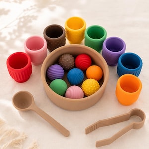 Può includere: Un set di giocattoli sensoriali colorati, tra cui palline strutturate, tazze, un cucchiaio e delle pinze. Le palline sono in vari colori e trame, e le tazze sono in tonalità di rosso, giallo, verde, viola, blu e arancione. I giocattoli sono disposti su una superficie neutra.