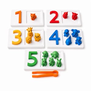 May include: Educational counting set featuring white trays with numbers 1-5 in various colors. Each tray has a corresponding number of animal figurines, including a fox, bears, giraffe, owl, lion, elephants, turtles, and bunnies. An orange tweezer is also included.