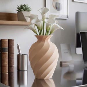 Op de afbeelding: Een lichtbeige vaas met een spiraalvormige, geribbelde textuur met een boeket witte calla lelies. De vaas heeft een smalle hals die aan de bovenkant iets uitloopt en de bloemen hebben gladde, trompetvormige bloemblaadjes met groene stelen.