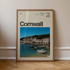 May include: A framed wall art print featuring a photograph of a coastal village in Cornwall, England. The print includes the text Cornwall, establishment date 1337, United Kingdom, and a list of regional landmarks, all set against a light beige background.