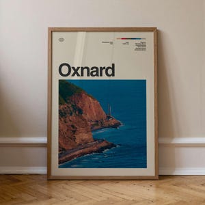 May include: A framed art print featuring the word "Oxnard" in bold black letters above a scenic coastal view. The image shows a reddish-brown cliff, a road, and the blue ocean. The print includes additional text and a color bar.