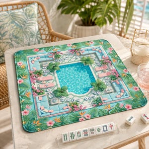May include: A square mahjong mat featuring a vibrant tropical pool scene with light blue lounge furniture, lush green palm leaves, and pink hibiscus flowers. The mat is displayed on a table with a rack of white mahjong tiles in the foreground.