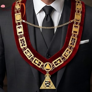 May include: A formal gold-colored chain collar with a red velvet band, adorned with geometric and symbolic designs. The collar is worn over a black suit and white shirt, with a black tie. A triangular pendant hangs from the collar.