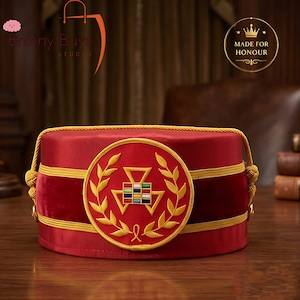 May include: A cylindrical red hat featuring a prominent circular emblem with a gold embroidered laurel wreath surrounding a multi-colored square grid and triangle design. The hat is accented with horizontal gold cord trim and side knots.