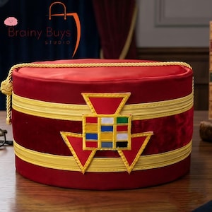 May include: A cylindrical red velvet hat featuring two horizontal gold braided bands and a gold cord trim along the top edge. A central embroidered emblem displays a grid of small colored squares in red, white, green, blue, and black.