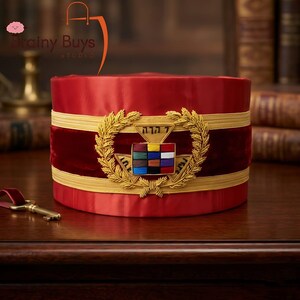 May include: A cylindrical red hat featuring a central gold embroidered wreath surrounding a rectangular emblem with twelve colored squares and Hebrew text. The hat is accented with horizontal gold bands and a deep red velvet middle section.
