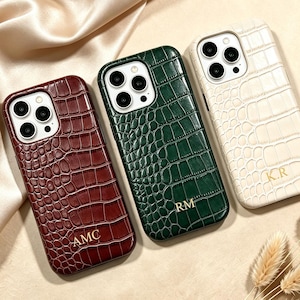 Puede incluir: Tres fundas para smartphone con textura de cocodrilo en color burdeos, verde oscuro y crema. Cada funda tiene un recorte para la cámara e iniciales doradas en relieve: AMC en la funda burdeos, RM en la verde y K.R en la funda de color crema.