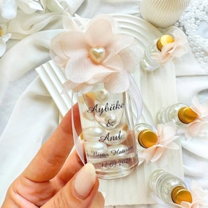 May include: A clear glass bottle filled with white pearls, topped with a pale pink fabric flower and a heart-shaped bead. The bottle features black printed text that reads Aybüke & Anıl, Nişan Hatırası, and the date 12.09.2024 in a script font.