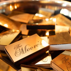 May include: A collection of rectangular matchboxes with a reflective gold metallic finish. Each box features the text The Moore's printed in a black cursive script on the top surface. The matchboxes are stacked together inside a round container.