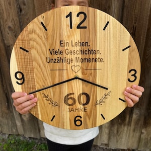 Op de afbeelding: Een ronde houten klok met zwarte cijfers en wijzers. De wijzerplaat toont de tekst "Ein Leben. Viele Geschichten. Unzählige Momente." en "60 Jahre." De klok wordt tegen een houten achtergrond gehouden.