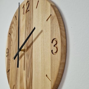 Könnte beinhalten: Eine runde Holzuhr mit natürlicher Maserung. Die Ziffern 12, 3, 6 und 9 sind in das Holz geschnitzt, mit Strichen für die anderen Stunden. Schwarze Zeiger zeigen die Uhrzeit an.