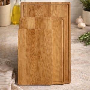 Può includere: Tre taglieri in legno di diverse dimensioni, disposti verticalmente. I taglieri sono di colore marrone chiaro con venature del legno visibili. Il tagliere più grande ha un bordo rientrante. Arredamento per cucina e sala da pranzo.