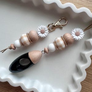May include: Two keychains featuring a sequence of wooden, white silicone, and beige gingham patterned beads. Each keychain includes a white daisy-shaped charm and a gold-toned metal lobster clasp. One keychain is attached to a black teardrop-shaped pendant.