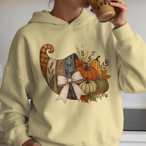 May include: A pale yellow hooded sweatshirt featuring a graphic print of a cornucopia filled with pumpkins, wheat stalks, and autumn leaves. The cornucopia is decorated with a large white bow and displays a plaid and leaf pattern on its surface.