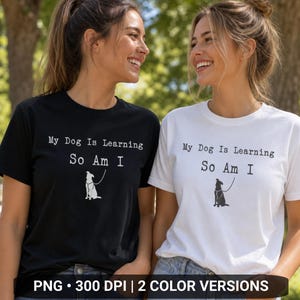 Puede incluir: Una camiseta negra y una camiseta blanca, cada una con el texto "My Dog Is Learning So Am I" en una fuente estilo máquina de escribir sobre un gráfico de un perro con correa. La camiseta negra tiene un gráfico blanco y la blanca uno negro.