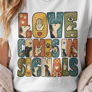 Könnte beinhalten: Ein weißes kurzärmeliges T-Shirt mit einem bunten Grafikdruck und dem Text LOVE COMES IN SIGNALS. Das Design enthält Illustrationen von Hunden, Handgesten, Pfotenabdrücken, Herzen und Augen in einem retro-inspirierten Raster.