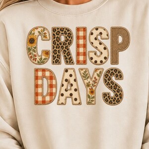 May include: A cream-colored crewneck sweatshirt featuring the text CRISP DAYS printed in large block letters. Each letter displays a unique pattern, including sunflowers, leopard print, orange and white gingham, polka dots, and brown burlap.
