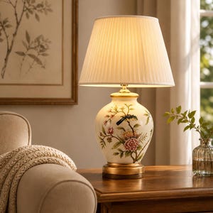 Puede incluir: Una lámpara de mesa de cerámica color crema con un diseño floral y una pantalla plisada. La lámpara tiene una base y detalles dorados. El diseño floral incluye flores rosas y amarillas con hojas verdes. La lámpara está sobre una mesa de madera.