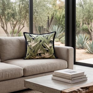 Peut inclure: Un coussin carré orné d'une bordure noire et d'un motif de feuilles tropicales vertes et blanches. Il est posé sur un canapé beige. Une table basse en béton avec des livres est au premier plan.