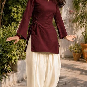 May include: A deep maroon long-sleeved tunic featuring a square neckline and a side-tie closure with flared sleeves, paired with loose-fitting off-white trousers that have a gathered, pleated design at the waist and ankles, worn with tan flats.