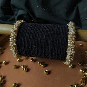 May include: Close-up of gold bangles adorned with small white pearls. The bangles are displayed on a black fabric roll, with scattered gold beads on a brown surface. The jewelry has a classic and elegant design.