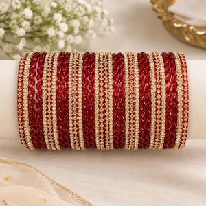 May include: A set of deep red bangles featuring a mix of smooth, wavy-textured bands and gold-toned bands encrusted with small, clear sparkling stones. The collection is arranged in a repeating pattern of red and gold, creating a layered look.