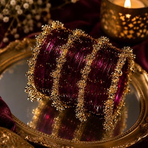 May include: A collection of deep red bangles with gold detailing. The bangles are stacked, displaying a rich, textured look. The gold accents have intricate patterns, enhancing the luxurious feel of the jewellery.