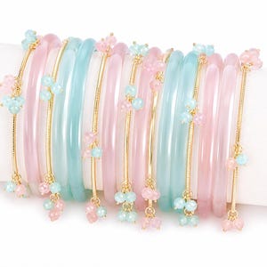 May include: A selection of pastel-coloured bangles. The bracelets are in shades of pink and teal, with gold-coloured chains and clusters of pink and teal beads. The bangles are stacked together on a white surface.
