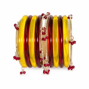 May include: A collection of vibrant bangles in yellow and red hues. The bangles are embellished with delicate gold chains and clusters of small red beads. The jewellery is displayed against a white backdrop.