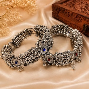 May include: Two silver-toned bangles featuring a textured, beaded surface and circular medallions. Each medallion is inlaid with a small, round blue or red stone. The bangles have a metallic finish and include small dangling charms along the edges.