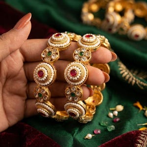 Puede incluir: Dos brazaletes dorados con un patrón repetitivo de motivos circulares. Cada brazalete está adornado con pequeñas cuentas blancas, piedras rojas centrales y piedras verdes más pequeñas engastadas en un marco metálico detallado.