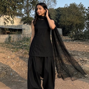 May include: A solid black sleeveless tunic paired with matching pleated loose-fitting trousers and a sheer black scarf. The ensemble features a simple round neckline and a consistent monochromatic colour scheme throughout the entire three-piece outfit.