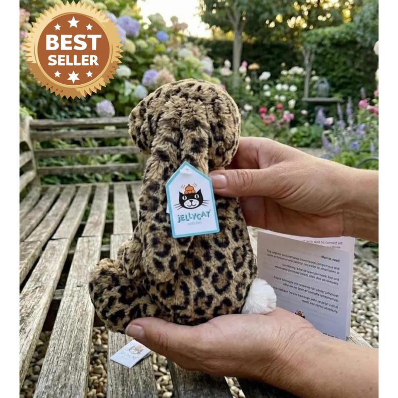 May include: A soft leopard-print toy animal with a white tail, held in hands. The tag reads "Jellycat". A "Best Seller" sticker is in the upper left corner. The background shows a wooden bench and a garden.