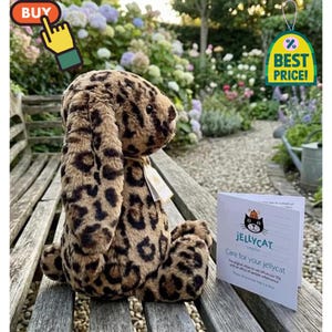May include: A soft toy rabbit with a leopard print pattern, sitting on a wooden bench. The toy has long, floppy ears and a tag attached. A sign that says "Best Price!" is in the background. A Jellycat card is also visible.