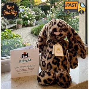 May include: A soft toy leopard-print bunny rabbit with long ears and a tag that reads "JELLYCAT." The bunny is next to a card that says "Care for your jellycat." The background includes a garden and the words "Best Quality" and "FAST."