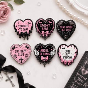 Op de afbeelding: Zes hartvormige emaille pins in zwart en roze met strikken, kruisen en teksten zoals Too Cute To Care, Spooky Babe, Dead Inside But Cute, Cry Baby Club, Angel Of Chaos en Cute But Psycho met druipende details.