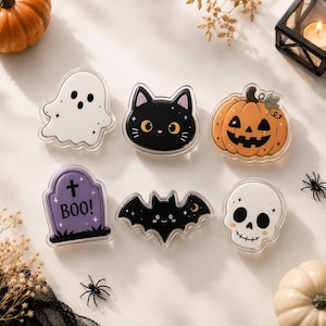 May include: Six acrylic Halloween-themed charms featuring a white ghost, a black cat face, an orange jack-o'-lantern, a purple tombstone with the text BOO, a black bat, and a white skull. Each piece has a clear border and a smooth, glossy finish.