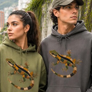 Op de afbeelding: Twee mensen dragen olijfgroene en antracietgrijze hoodies met een hagedissenontwerp. De hoodies hebben een kangoeroezak en een capuchon met trekkoord. De hagedissenafbeelding heeft groene, gele en zwarte vlekken. De achtergrond bevat een muur met de tekst "PAISA STUDIO".