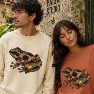 Può includere: Due felpe, una color crema e una color ruggine, ciascuna con una grafica a forma di rana. Il design della rana incorpora toppe verdi, nere e beige. Le felpe sono a maniche lunghe e hanno una vestibilità ampia. Il testo "PAISA STUDIO" è visibile sul muro dietro di loro.