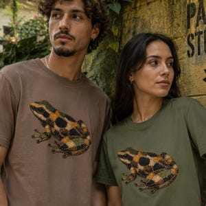 Könnte beinhalten: Zwei Personen tragen T-Shirts mit Froschgrafiken. Eines ist braun, das andere olivgrün. Das Froschdesign hat schwarze, orange und grüne Muster. Der Hintergrund zeigt eine gelbe Wand mit dem Text "PAISA STUDIO" und eine Pflanze.
