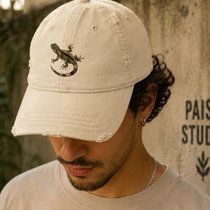 Può includere: Un cappellino da baseball beige con bordo usurato, con un disegno di lucertola. Il cappellino ha una visiera curva e un design a sei pannelli. Il testo "PAISA STUDIO" è stampato sul muro dietro il cappellino.