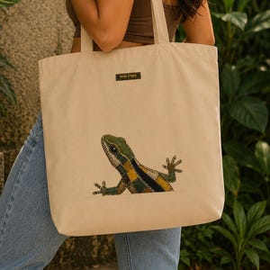 Op de afbeelding: Beige canvas draagtas met een illustratie van een hagedis in groen, geel en zwart. De tas heeft lange hengsels en een klein label met "PAISA STUDIO". De achtergrond toont groene bladeren en een stenen muur met de tekst "PAISA STUDIO".