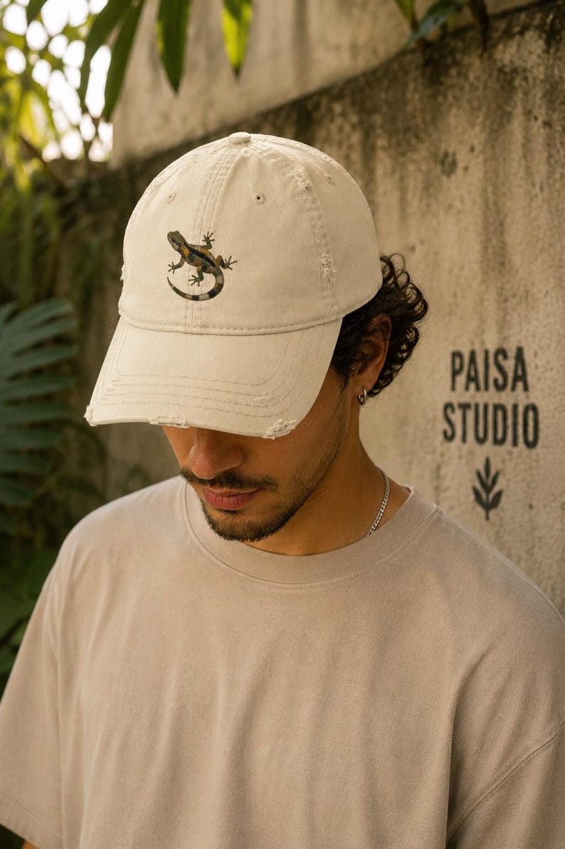May include: A tan baseball cap with a distressed brim features a lizard graphic. The cap has a curved bill and a six-panel design. The text "PAISA STUDIO" is printed on the wall behind the cap.