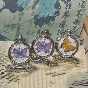 May include: Three silver-toned pocket watches with white faces and Roman numerals. Each watch features a butterfly motif on the dial: two with purple butterflies and one with a yellow butterfly. The watch covers are decorated with small floral accents.