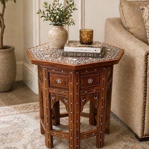 May include: A dark brown, hexagonal side table with inlaid white floral designs. The table has two small drawers and decorative legs. On top are two books, a vase with greenery, and a textured glass. The table is approximately 61 cm tall.