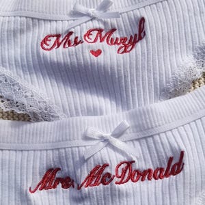 May include: Two pairs of white ribbed underwear featuring lace trim and a small white bow at the center waist. One pair is embroidered with the red text Mrs. Muzyl and a small red heart, while the other features the red embroidered text Mrs. McDonald.