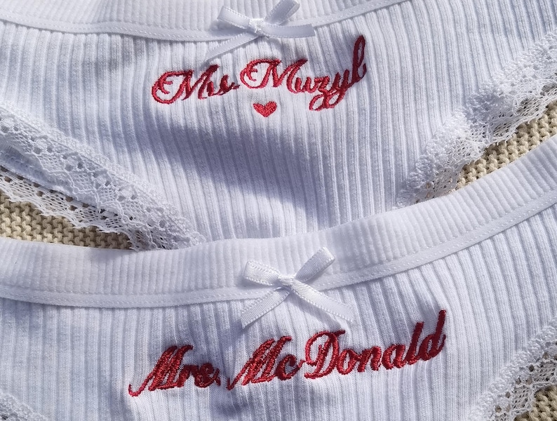May include: Two pairs of white ribbed underwear featuring lace trim and a small white bow at the center waist. One pair is embroidered with the red text Mrs. Muzyl and a small red heart, while the other features the red embroidered text Mrs. McDonald.
