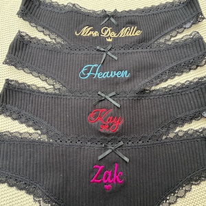 May include: Four pairs of black ribbed underwear featuring black lace trim and a small black bow at the center. Each pair is embroidered with different text: Mrs. DeMille in gold, Heaven in light blue, Kay in red, and Zak in bright pink.
