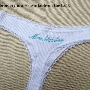 May include: A white ribbed thong featuring delicate lace trim along the edges and the text Mrs. Geisler embroidered in light blue thread on the front. The text Embroidery is also available on the back is visible above the garment.